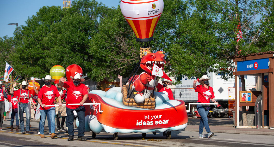 Volunteers in red UCalgary t-shirts push the UCalgary float, which has Rex on the front and a small hot air balloon on top. 
