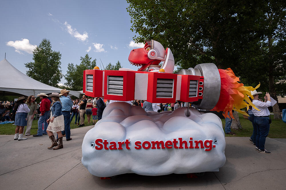 A parade float with a base shaped like a cloud with the words "Start something" on it. Above the cloud base is a spaceship shaped like the Start something logo being driven by Ucalgarys mascot, Rex, in space attire.