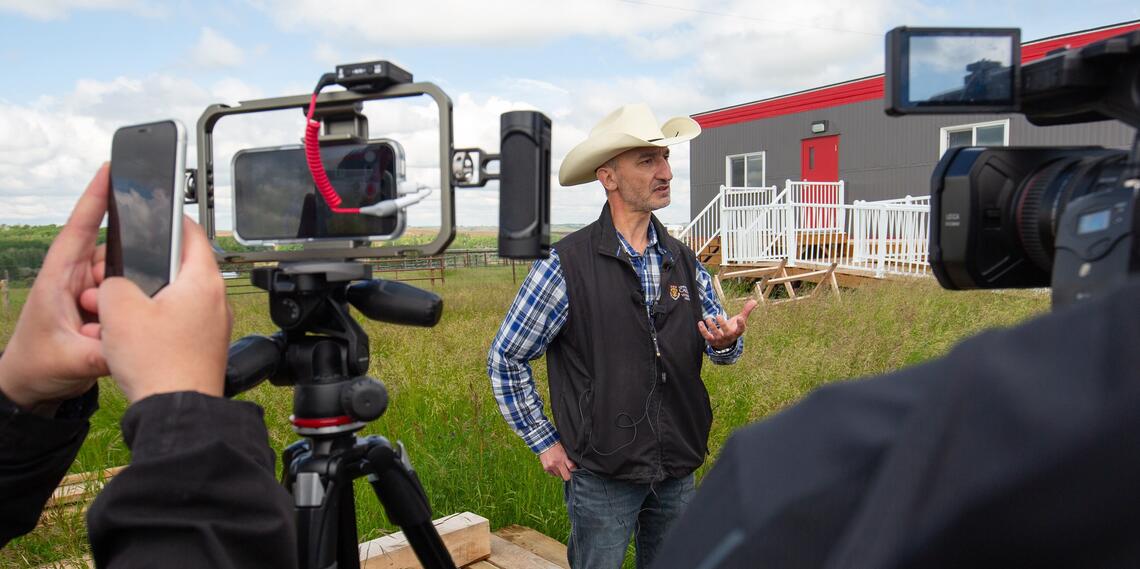 A man in a cowboy hat and UCalgary shirt talks to film cameras