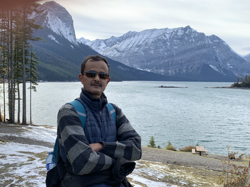 A man wearing sunglasses stands in front of lake with mountains in the background.