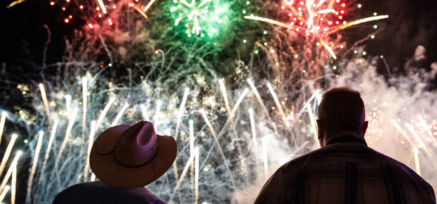 Two men stand silhouetted in front of red, green and white fireworks. One man wears a cowboy hat.