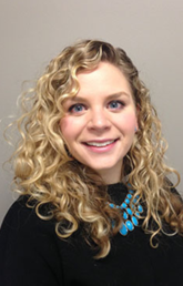 Alumna Audrey Taylor Bereznicki faces the camera and smiles. She is wearing a black shirt, has shoulder length curly hair, and stands against a beige background.