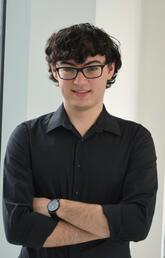 A young man with dark hair, glasses and wearing a dark button down shirt smiles at the camera with his arms crossed