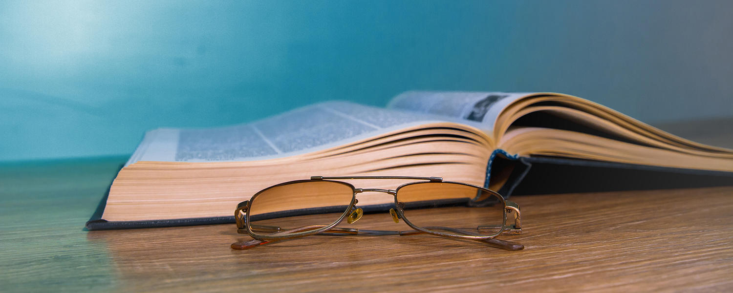 An open book sits on a wooden table next to a pair of reading glasses. 