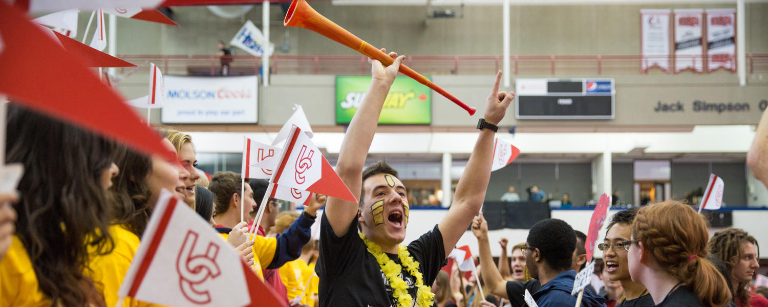 A crowd of students with UC flags. There is a young man in the center with his arms up cheering. He has Facepaint and is wearing a yellow Lei 