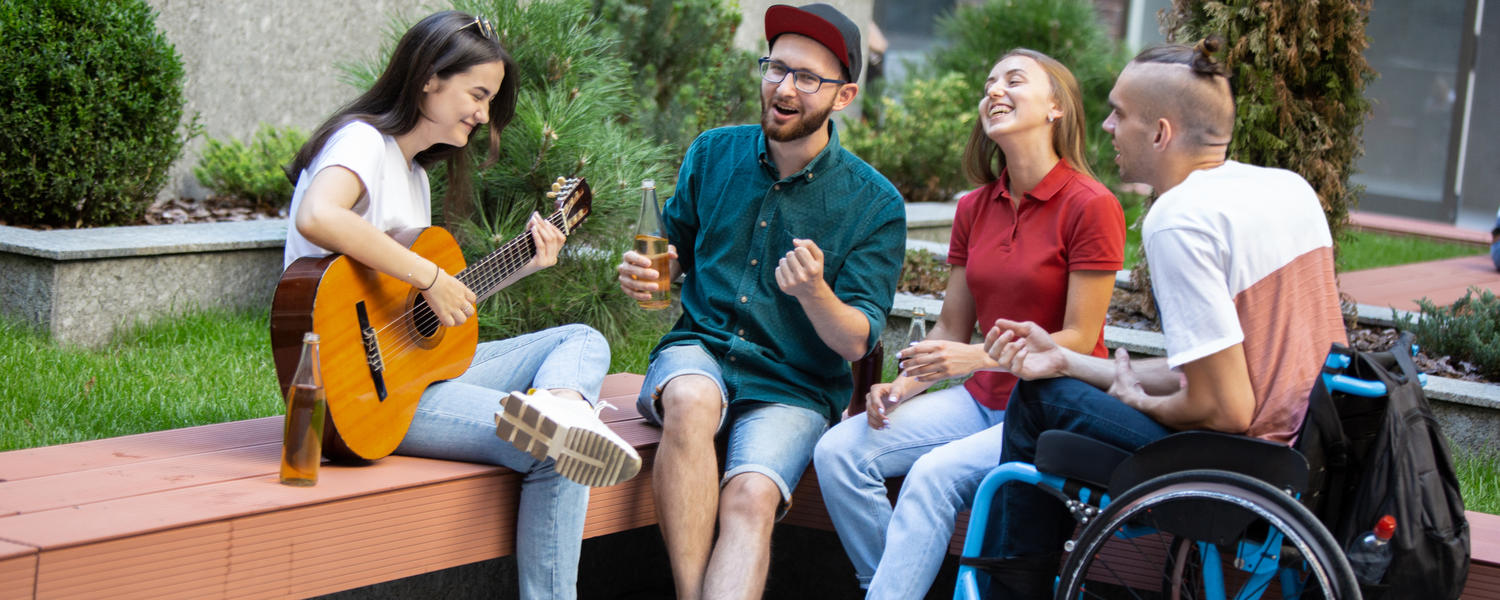 A group of two males and two woman are seated on a bench. One of the men are in a wheelchair. One of the women is playing guitar.