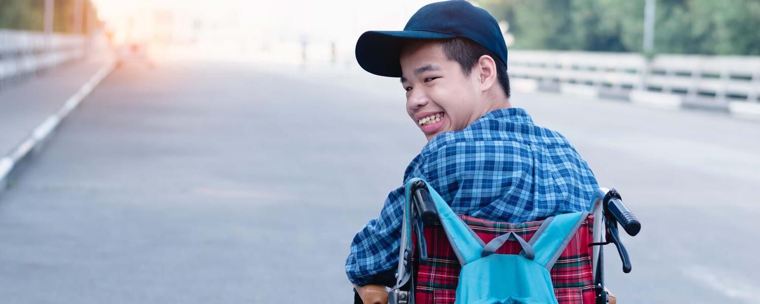 Smiling boy in blue cap and shirt with a blue backpack, and sitting in a wheelchair heading up a road
