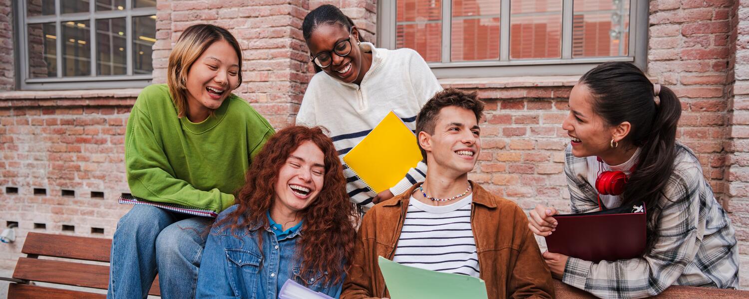 A photo of four young people, presumably students are seating and standing on a bench in front of a red brick wall. They are chatting and laughing with each other. They have duotangs and binders in their hands 