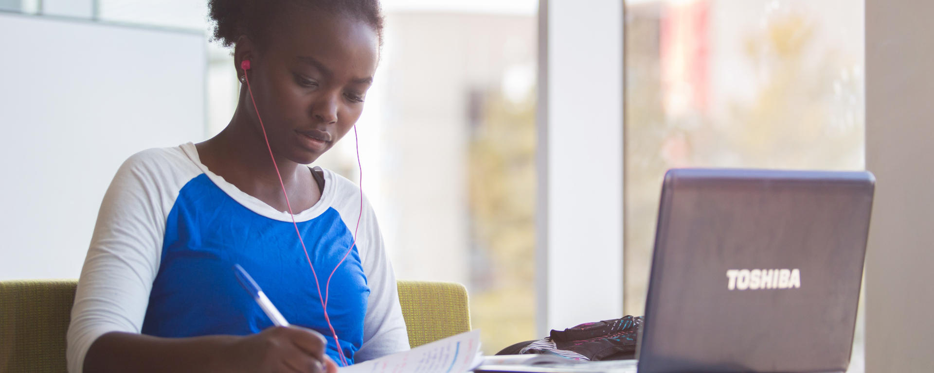 Woman is seated next to a bright window working on her laptop