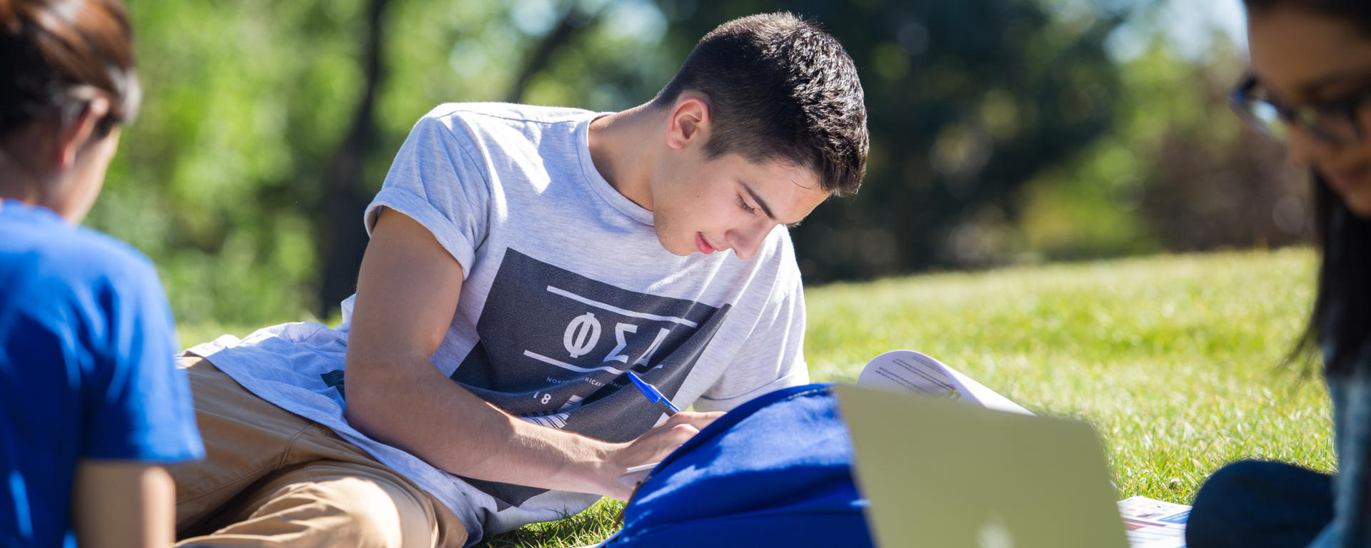 Three (presumably) students sit on green grass. There is a woman on the left in a blue shirt with her back to the camera, a young man with brown hair laying as he looks down at his book with pen in hand, and a woman wearing glasses on the right side of the screen looking down at her laptop
