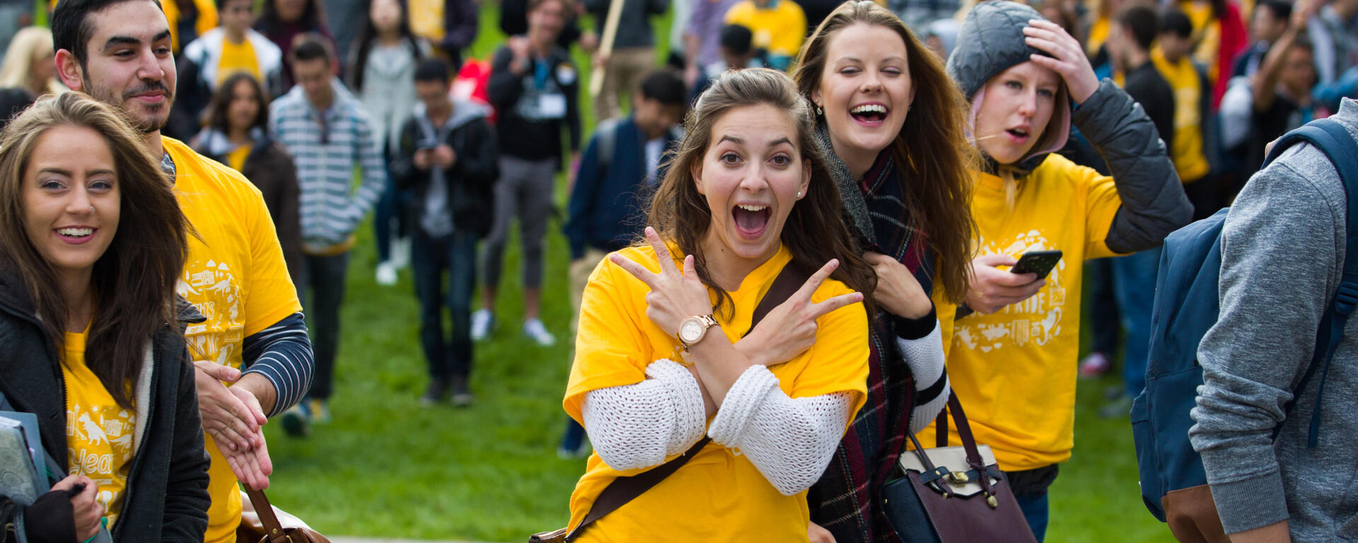 A crowd of students, one is looking at the camera giving two peace signs. Most are in yellow tee shirts 