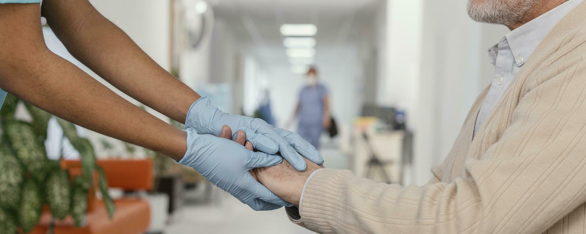 arms of a doctor wearing gloves coming into the frame from the left and holding the hands of an elderly white gentleman in a wheelchair. 