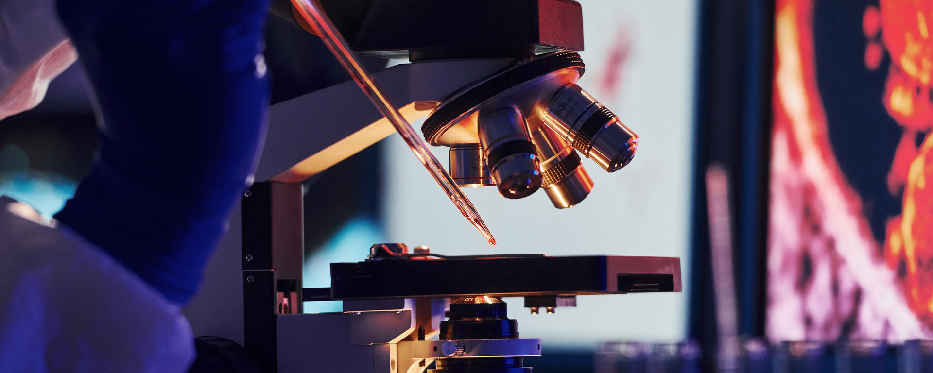 Gloved hands using a pipette to apply liquid to a slide under an illuminated microscope, with test tubes in the background.