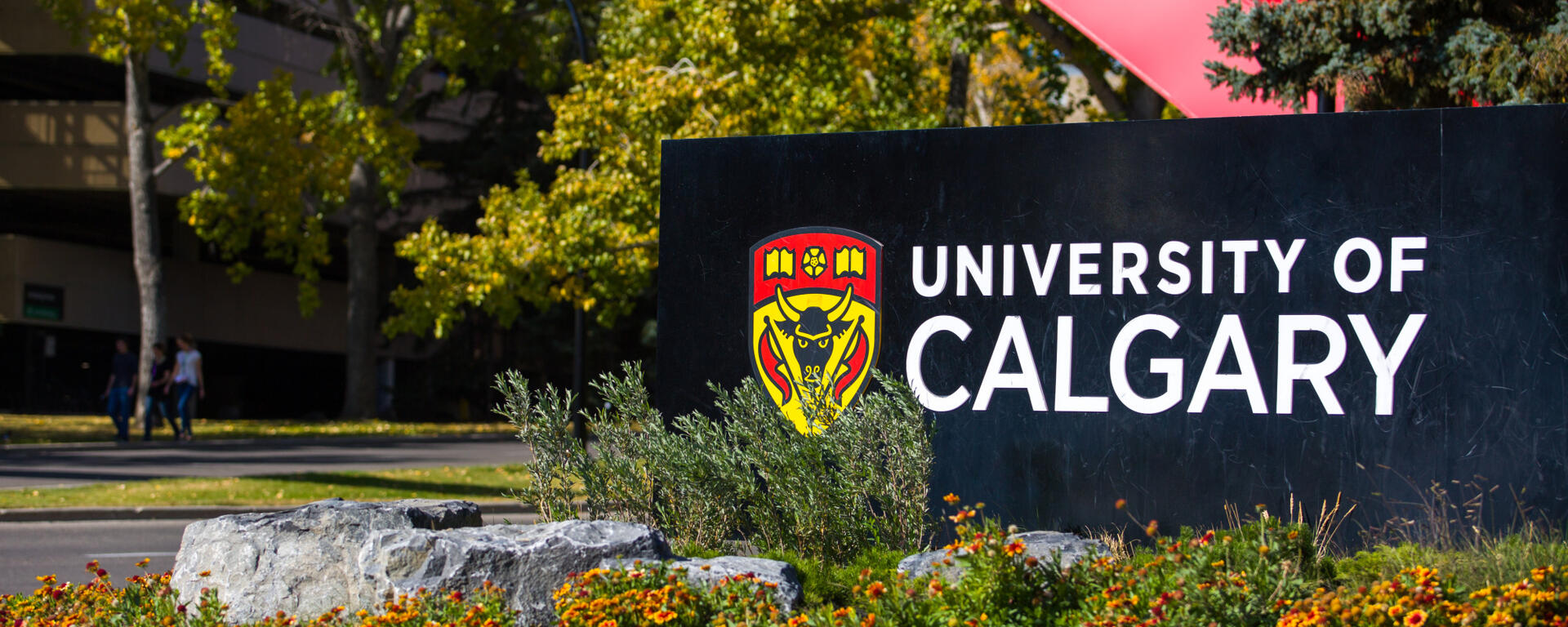 A photo of the U of C Sign at the front of campus, just below the two red arches. Below the sign is orangey red and yellow flowers. The trees in the background are green and yellow, indicated the fall season has just begun. 