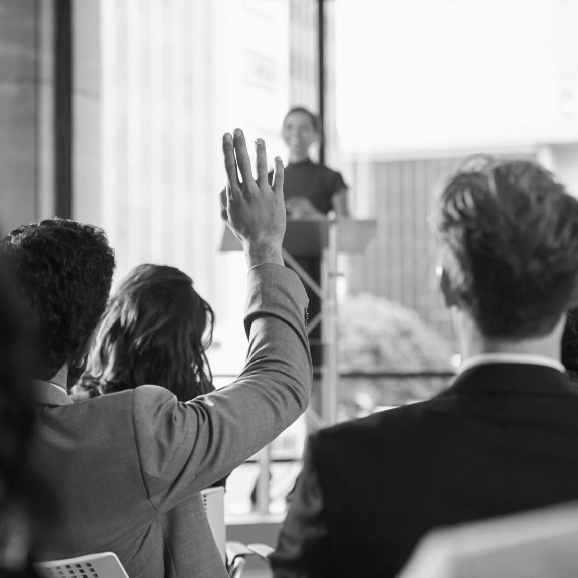Black and white photo of a crowd of people at a conference. One person in the audience has their hand up. The individual at the is blurred out and far away.