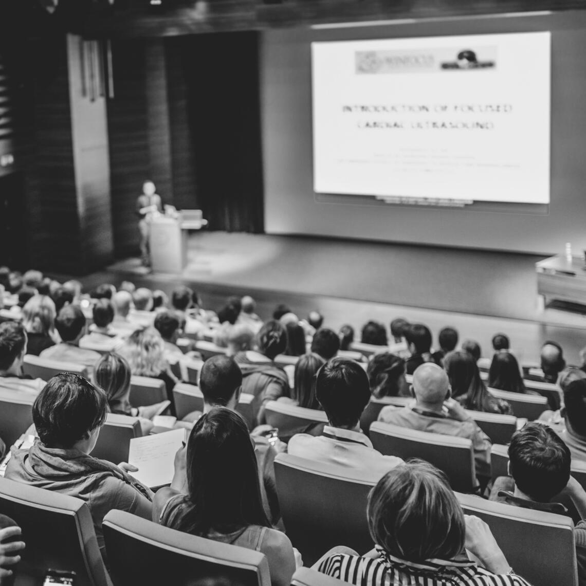 Black and white photo of a crowded conference. The photo is facing towards the presenter so you see the crowd from behind. The presenter screen is on stage and both the screen and podium are blurry