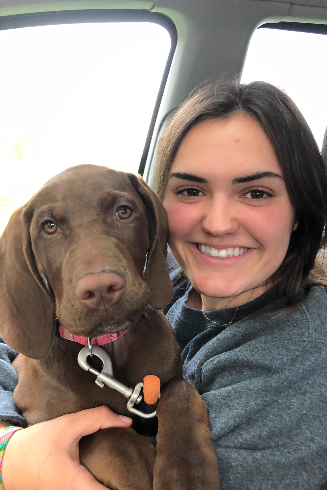 A photo of Maeve Butts smiling as she holds a chocolate lab puppy. Shje is wearing a grey hoodie with a black tee shirt underneath. Her hair is brown. 