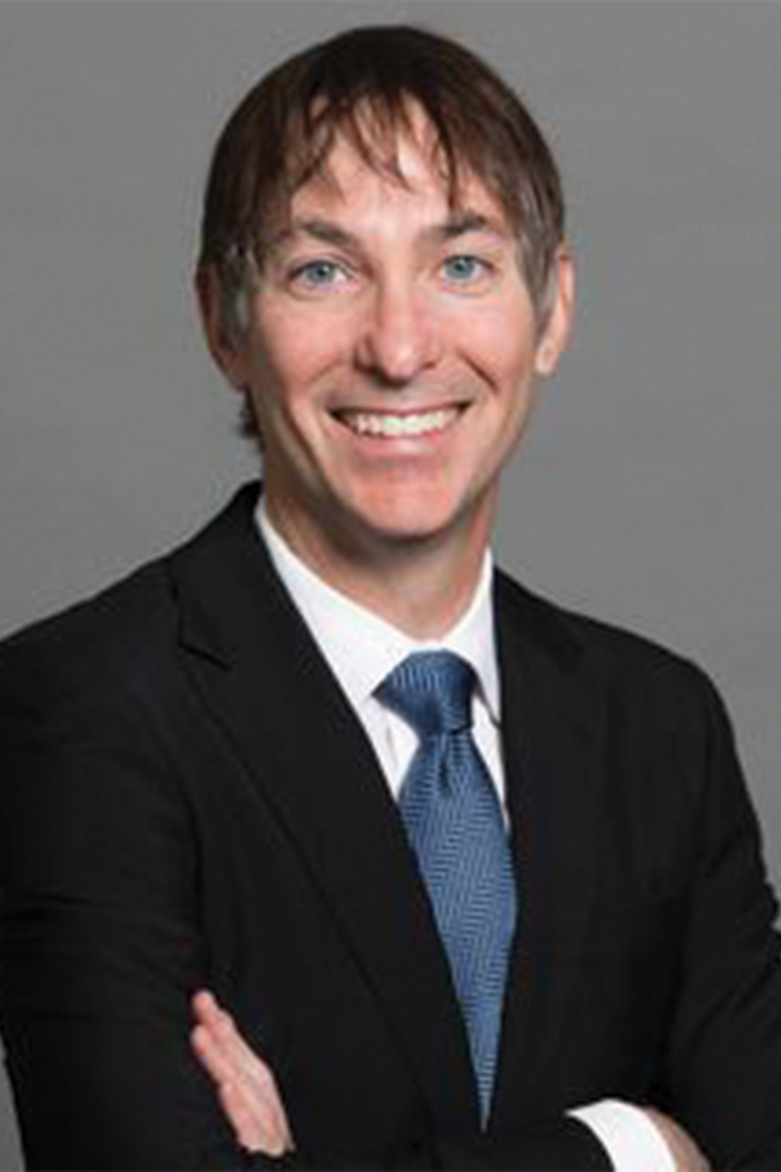 Professional head shot of Dr. Cello Tonelli. Wearing a white collared dress shirt, blue tie, black jacket and has his arms crossed.  