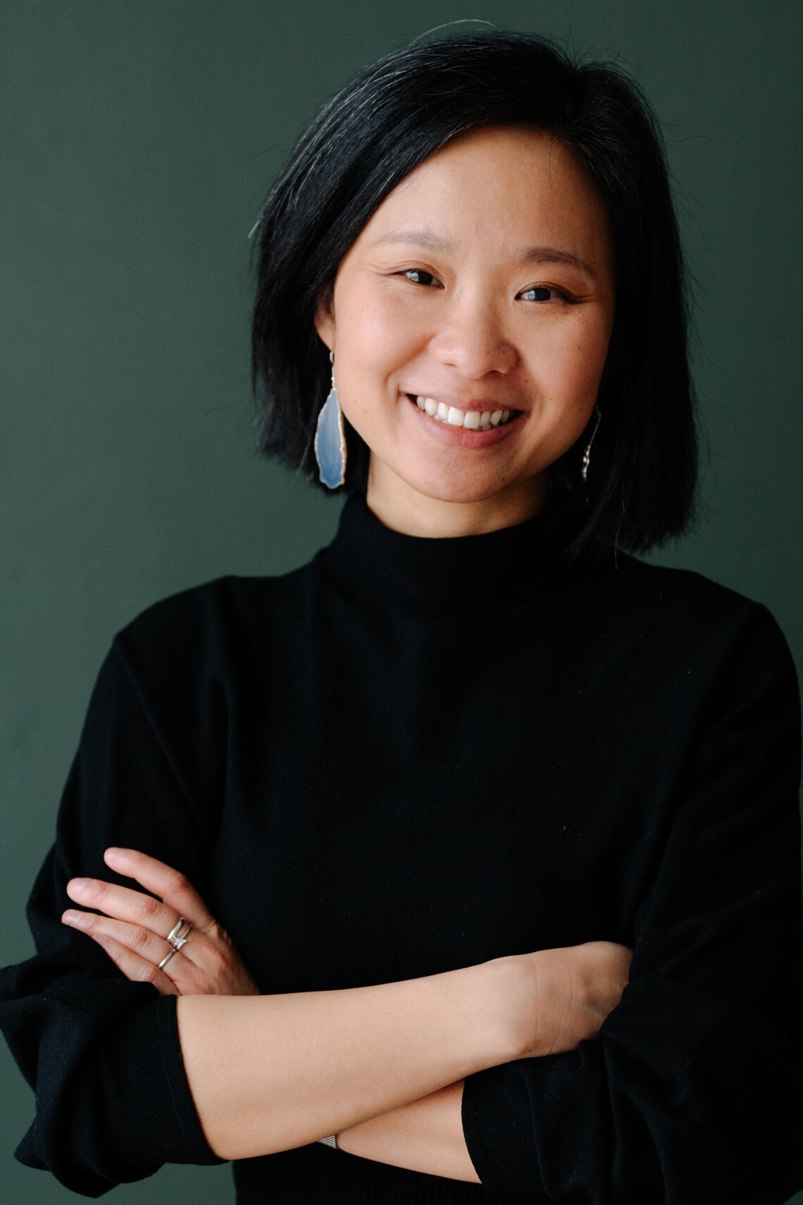 Professional photo of Dr. Helen Tam-Tham, standing with arms crossed, wearing a black sweater with pants. She is smiling, has shoulder length black hair with grey streaks. She has long earrings one. 