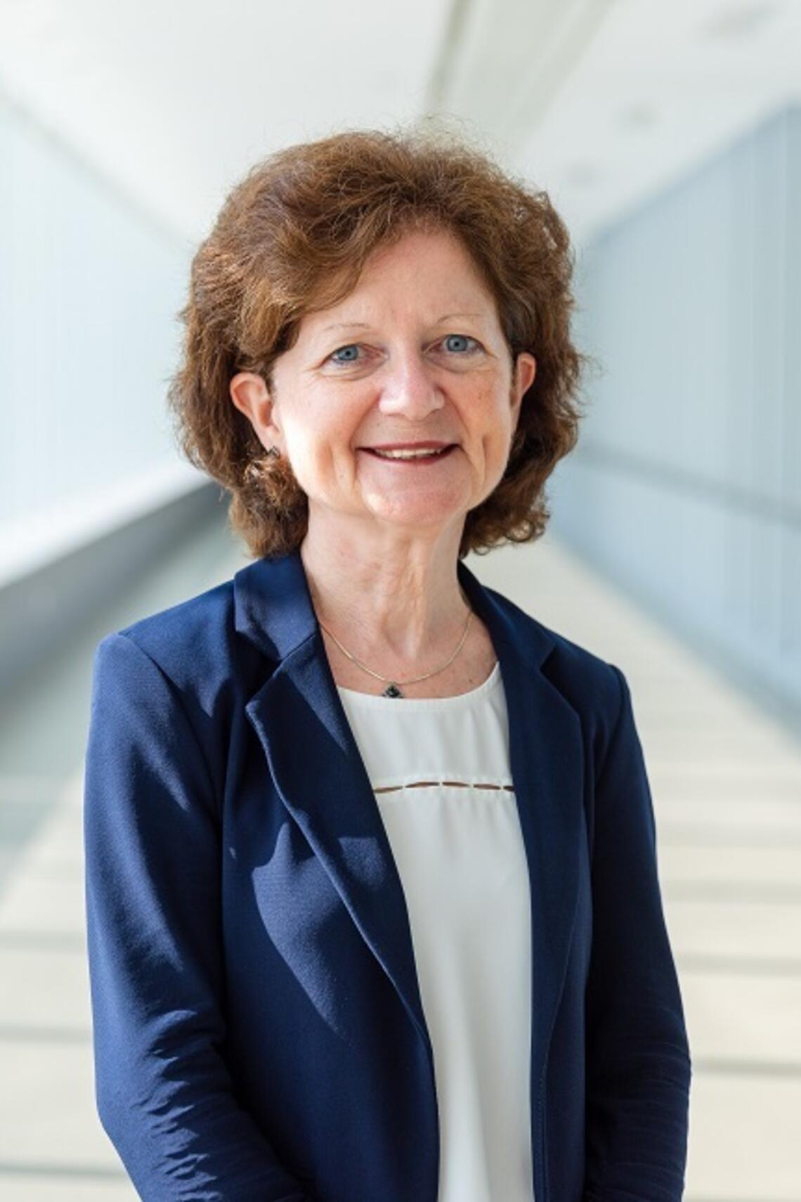 Professional photo of Dr. Brenda Hemmelgarn. Standing in a blurred hallway. Wearing a Navy blue jacket and white shirt. She has brown hair cut to just above the shoulders. 