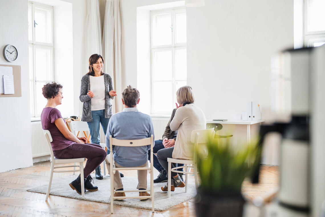 Photographof a woman standing and talk to four people sitting in chairs. Appears to be a group discussion or an educator speaking to learners.