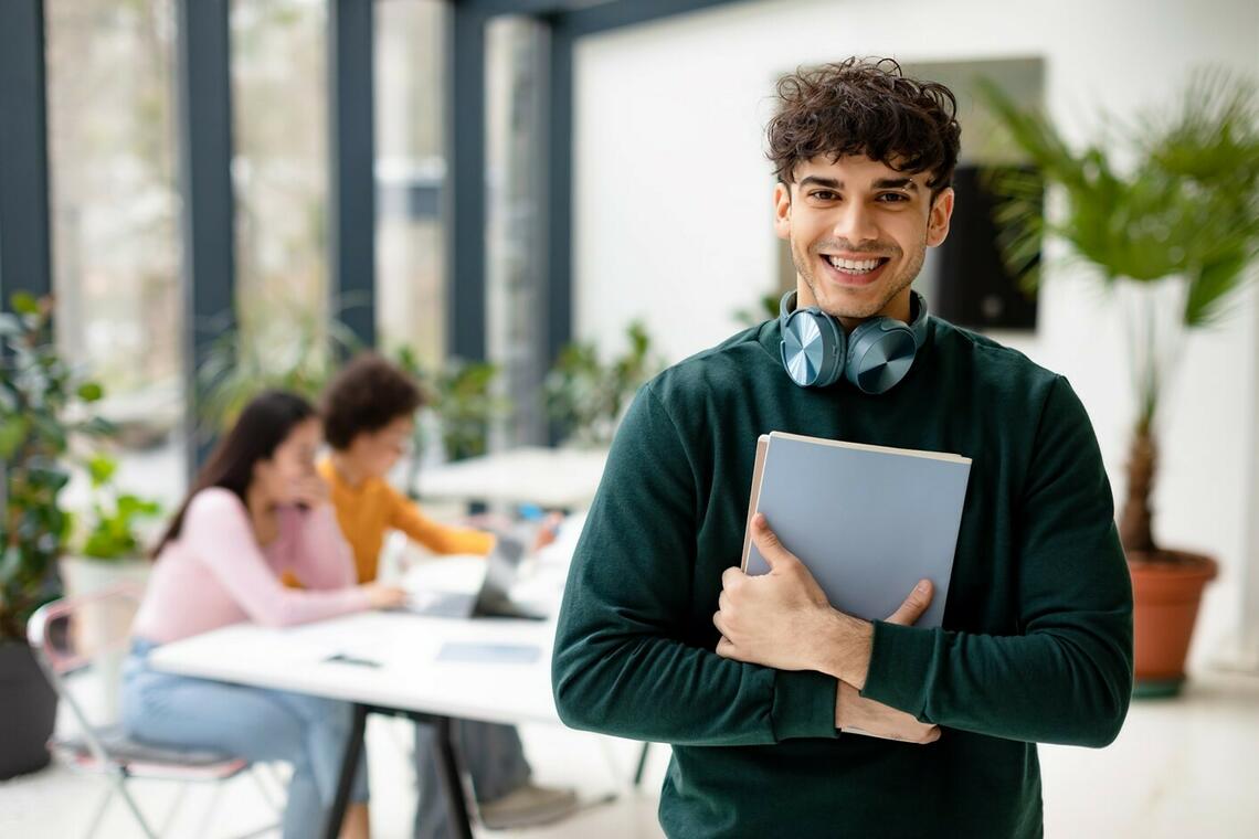Male college student holding books and smiling at camera while female classmates studying with laptop on the background in a  coworking space