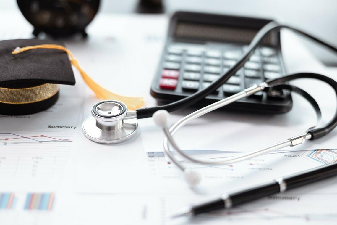 A graduation cap, stethoscope, calculator, pen, clock and papers placed on top of a table. 