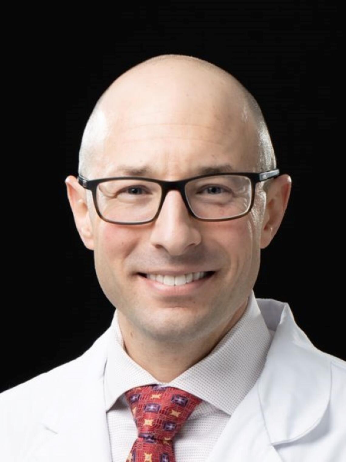 A photo of Nowell Fine, a smiling bald white man in his forties wearing a white coat, dress shirt and red patterned tie.