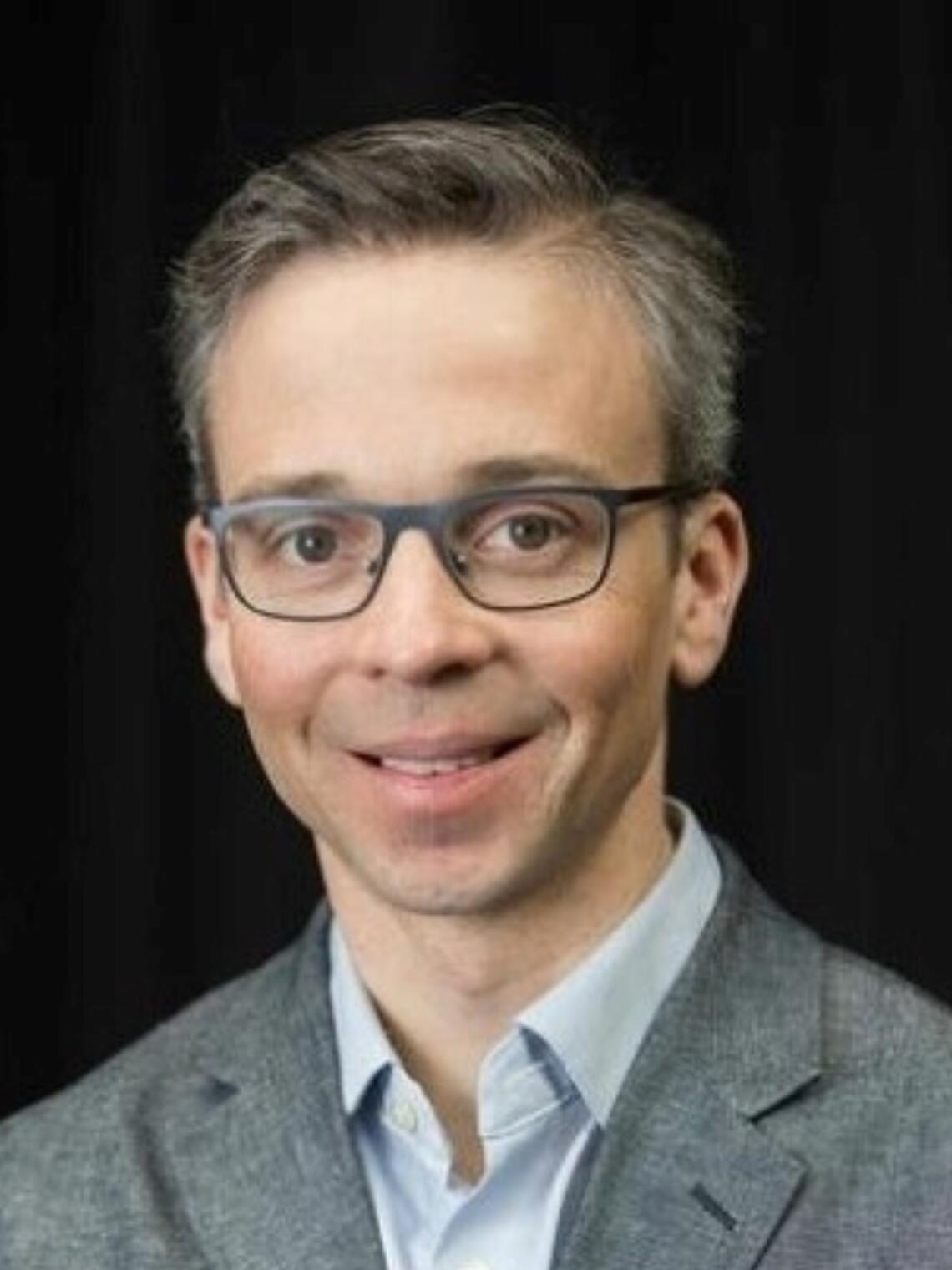 Photo of Dr. Stephen Wilton, a smiling white man in his forties with salt-and-pepper hair, wearing a black v-neck sweater and white dress shirt underneath.