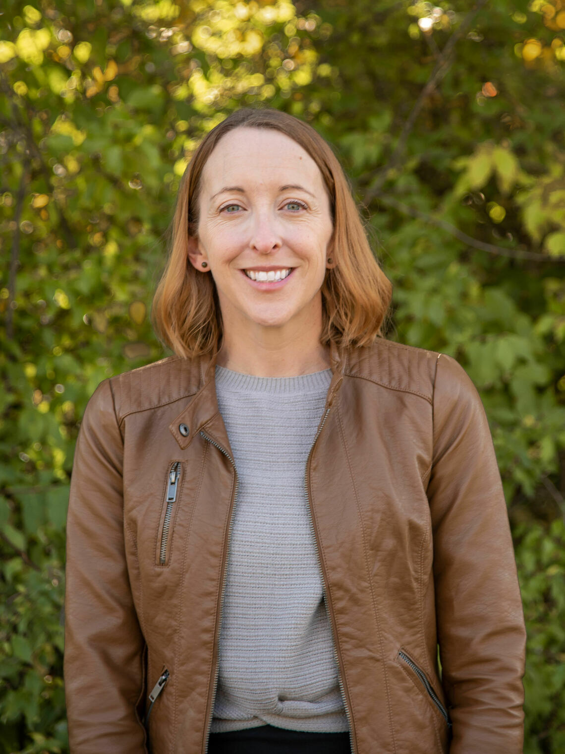Smiling woman with shoulder length light brown hair and wearing a brown jacket and grey sweater facing the camera and standing in front of trees