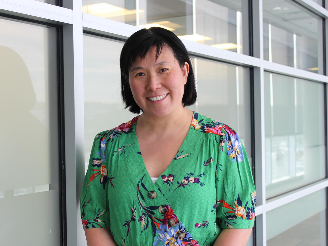 Photo of Dr. Pamela Chu. She is smiling, standing in front of a glass wall, wearing a green floral-print shirt.