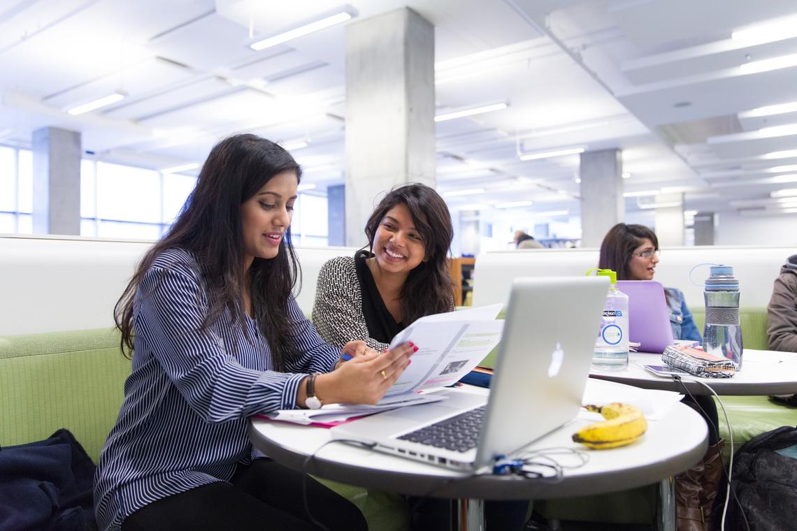 Two women are seated at a round table looking at a paper with a laptop in front of them and a banana