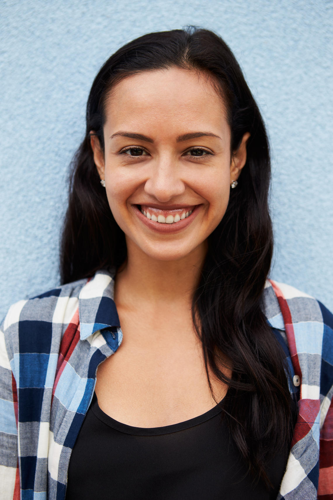 Portrait of a woman with long black hair. She is smiling and wearing a red/blue/white checkered flannel top with a black tank top underneath