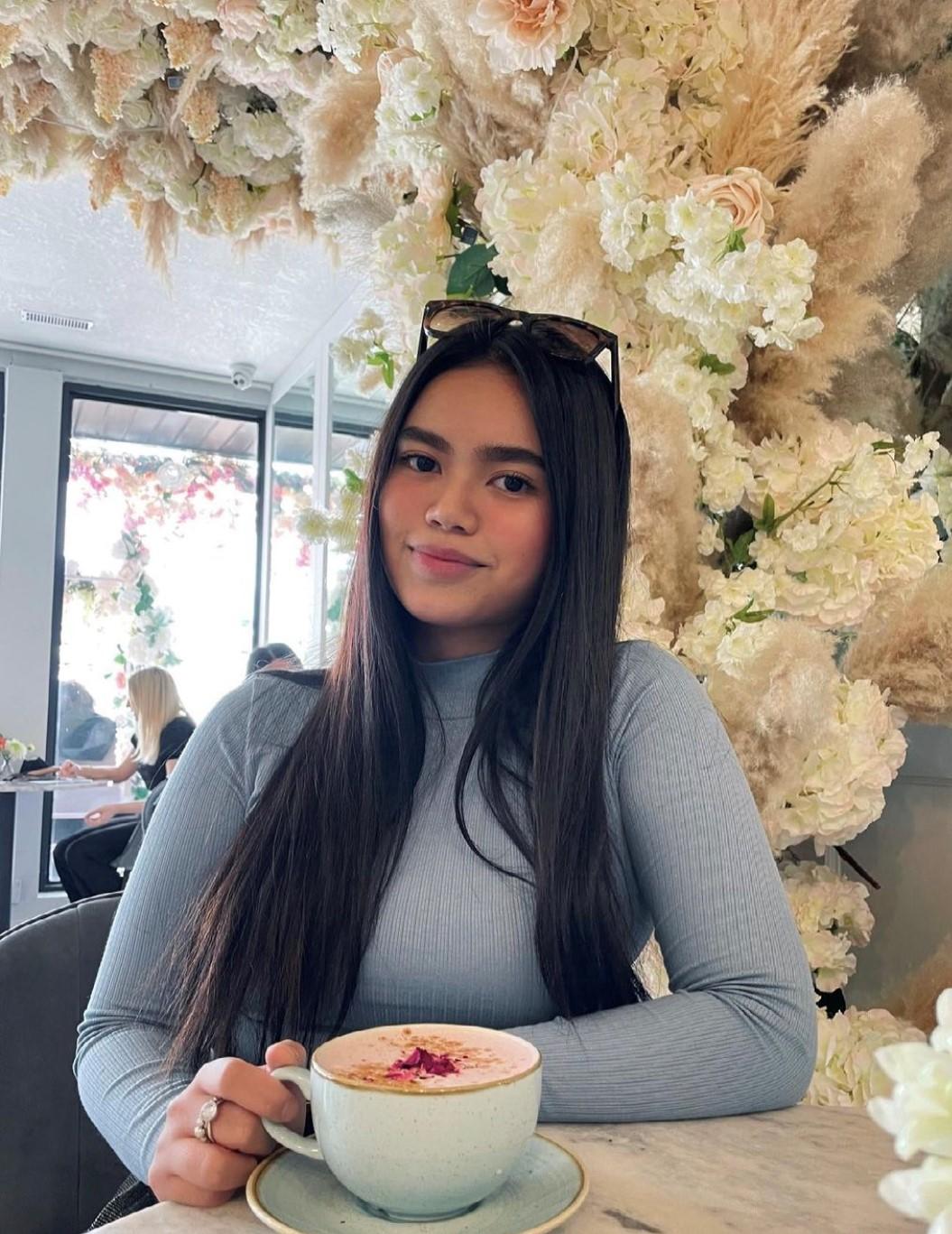 A portrait of Denise Banaag. She is seated with a coffee in front of a wall of white flowers