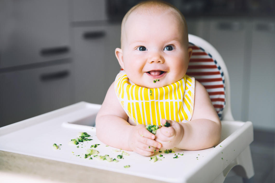 baby in highchair with yellow bib and green food