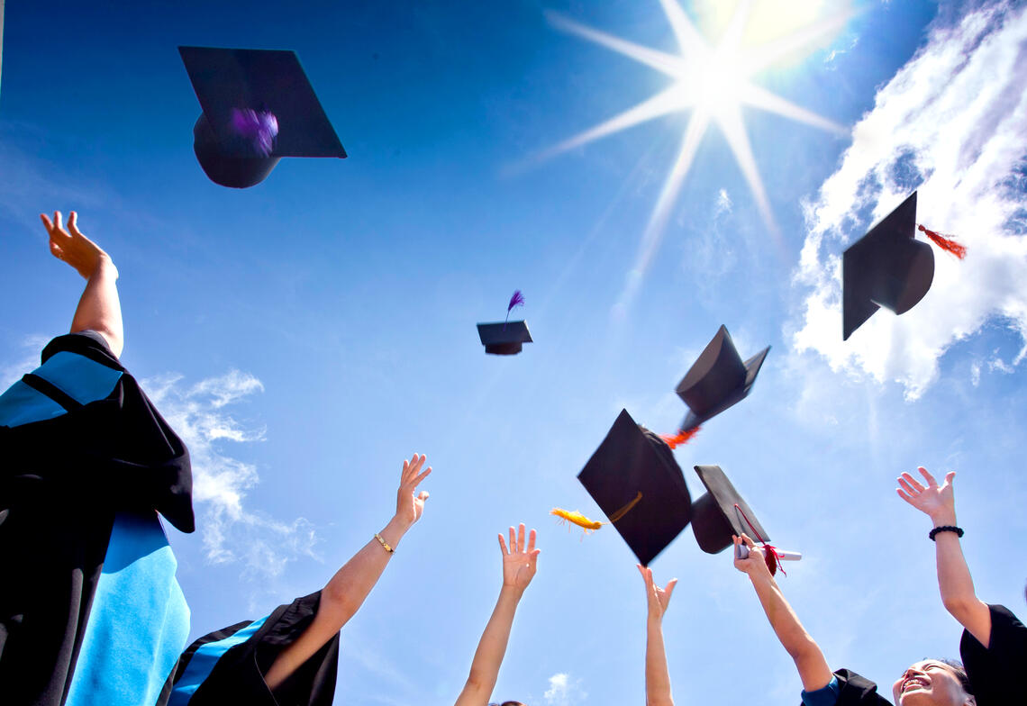 A photo from ground view up of people dressed in graduation gowns. They are throwing their graduation caps in celebration towards the blue sky and bright shining sun.