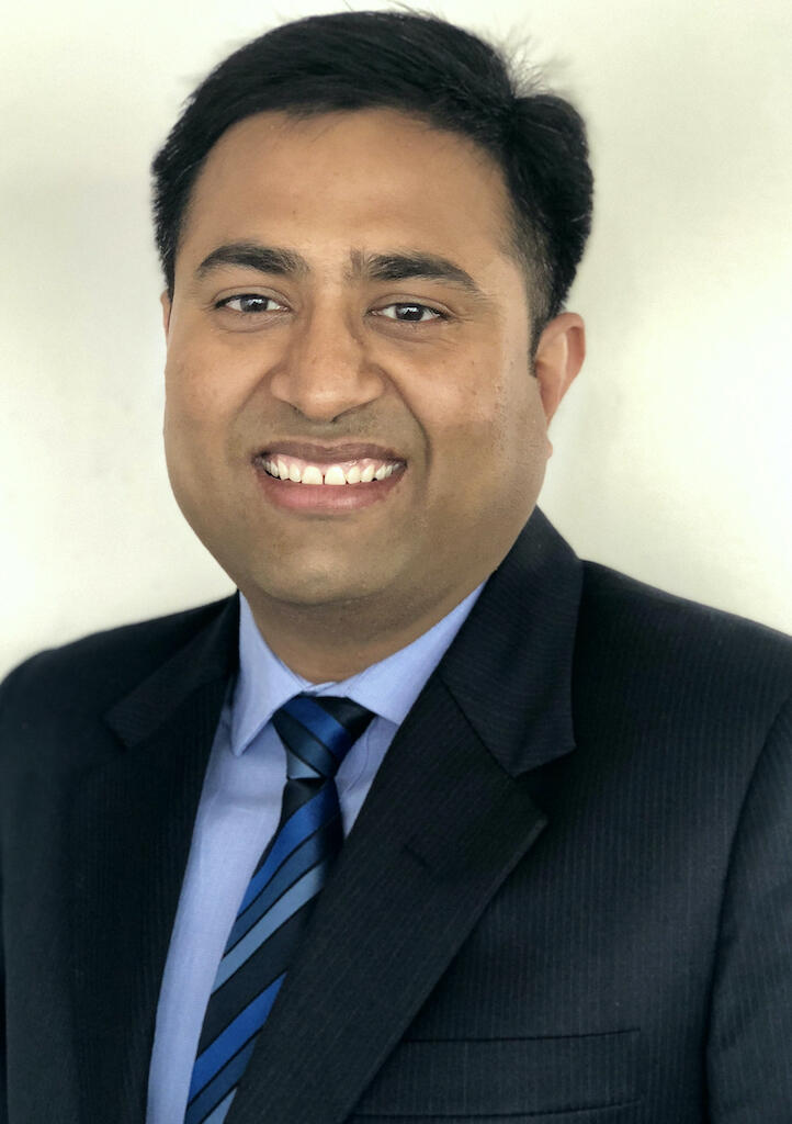 Photo of man of Indian descent wearing black blazer, blue collared shirt, blue striped tie