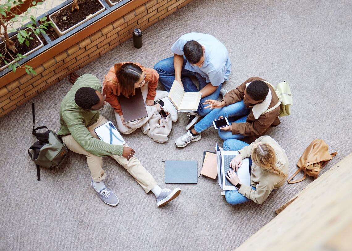 Birds eye view photo of 5 students having a meeting on the floor. There are books and laptops scattered on the floor. They look to be chatting and collaborating.