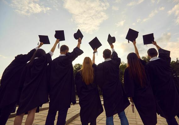 A photo of the backs of 7 students, all wearing their Graduation caps and gowns. They are all holding their caps towards the sky in celebration of graduating. The sky looks to be around sunset with a golden huge