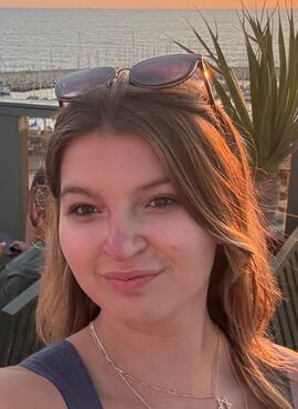 CRDSSA VP Events and Communications Andrea Huber. Andrea has mid length light brown hair. The photo is selfie style. She is smiling at sunset with a body of water behind her and sunglasses on top of her head