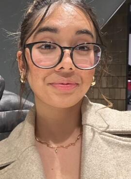 CRDSSA VP Administration Gunpreet Minhas. Gunpreet has dark brown hair in an updo. She is smiling in a selfie style photo. She is wearing black round glasses, an oat colored jacket, with a white top underneath. 