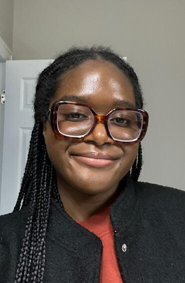 CRDSSA Junior Administration Ibukun Olaniyan. Ibukun has briaded long black hair. She is smiling in a selfie style photo with tortoise shell glasses. She is wearinng a black jacket overtop of a burnt orange top.