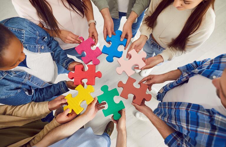 A birds eye view of seven people holding different colored puzzle pieces in a circle trying to piece them together 