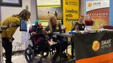 A young girl in a wheelchair demonstrating a switch adapted toy with her mom, brothers and occupational therapist watching