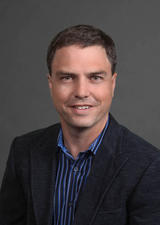 Dr. Sean Dukelow wears a black blazer with a royal blue collared shirt. He smiles at the camera for his headshot.