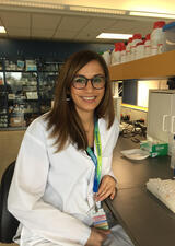 Dr. Cindy Barha wears a lab coat, nametag and glasses. She sits at a laboratory bench and smiles.