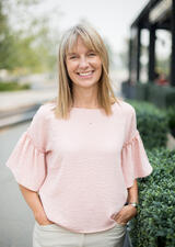 Dr. Sonja Wicklum stands outdoors facing the camera smiling. She is wearing a pink top with flowing sleeves and stonewashed khakis. Her hands are in her pockets. 