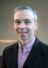 Dr. Stephen B. Wilton smiles at the camera. He is wearing a dark salmon dress shirt and charcoal suit jacket. 