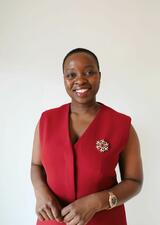 Dr. Nonsikelelo Mathe stands facing the camera, smiling. She is wearing a red short sleeved shirt with a white and red brooch on her left hand side, and a gold watch on her left wrist. 