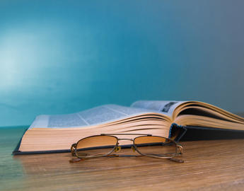 An open book sits on a wooden table next to a pair of reading glasses.